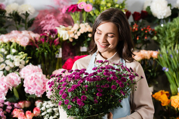 Front view of smiling female florist looking at purple chrysanthemums in vase near blurred racks of flowers on background