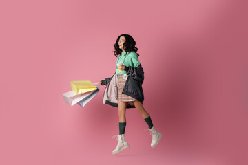  brunette young woman in casual winter outfit with shopping bags and coffee to go jumping on pink background