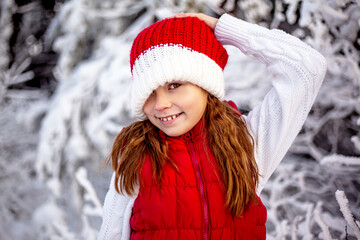 Happy girl playing with snow in frosty winter . Child dressed red christmas hat , red vest and white blouse, walking in the snowy nature