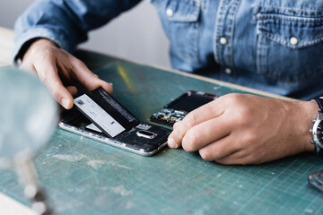 Cropped view of repairman putting battery in mobile phone on table with blurred magnifier on foreground