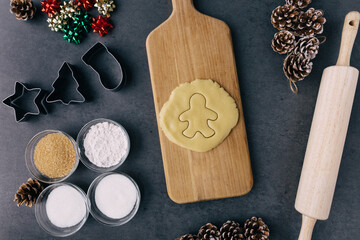 Cutting Christmas Ginger Bread Man Cookie In Raw Cookie Dough With Pine Cones, Holiday Ribbons, And Ingredients on Dark Slate Texture