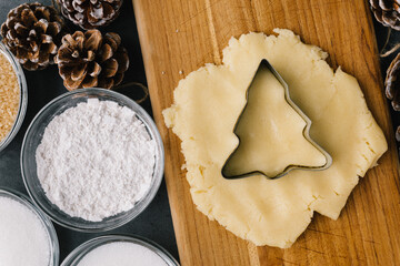 Cutting Holiday Christmas Tree Cookie In Raw Cookie Dough With Pine Cones And Ingredients on Slate