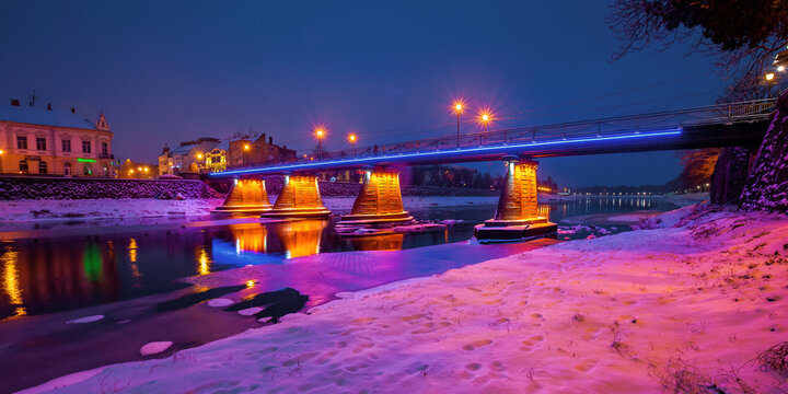 uzhhorod, ukraine - 26 DEC, 2016: old town on a christmas night. beautiful cityscape by the river. snow on the embankment. bridge and lanterns glowing in the distance