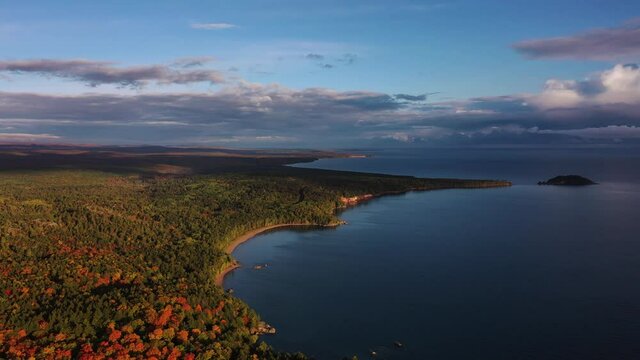 Beautiful aerial panning shot over the coastline of Lake Superior with tree covered forests below and clouds in the blue sky above.