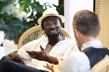 African American millionaire is talking about business with another Caucasian white man in  luxury tropical style hotel lobby - obrazy, fototapety, plakaty