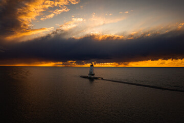 Beautiful aerial photograph of the Ludington Breakwater Lighthouse at the end of a concrete pier in front of a beautiful orange sunset sky as it reflects on the calm water of Lake Michigan.