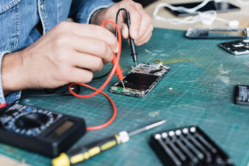 Cropped view of repairman holding multimeter sensors on disassembled part of mobile phone on blurred foreground