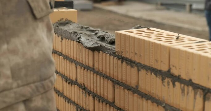 Bricklayer finishes brick row, fills last gap in brickwork, distributes mortar, takes out brick, stacks it, adjusts it with trowel according to level, removes excess mortar, construction site