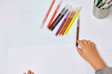 Naklejka premium Cute little boy with blond hair draws colored pencils at home. Draws at the white table. Close up of child's hands drawing at white paper