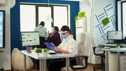 Obraz premium Teamworkers in new normal office making financial strategy wearing protective mask. Employees with visors working in corporate company workspace respecting social distance analysing data and graphs.