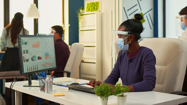 Black woman with protection face masks working on computer in workplace during pandemic. Multiethnic team in new normal business financial office checking reports, analysing datas looking at desktop