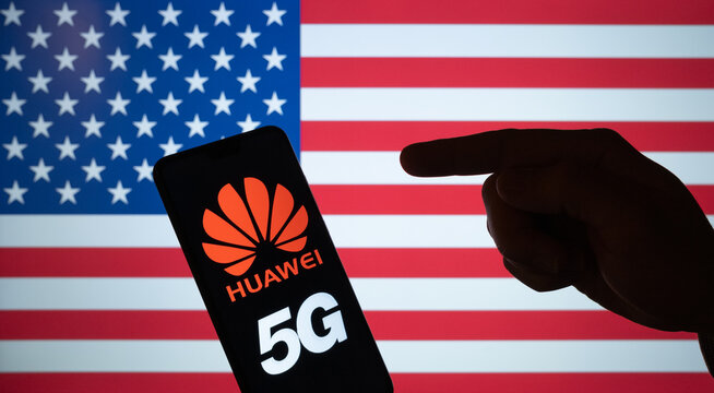 Stone /UK - July 16 2020: Huawei logo on smartphone silhouette and hand pointing at it. The US blurred flag on the background screen. NOT A MONTAGE, real photo.