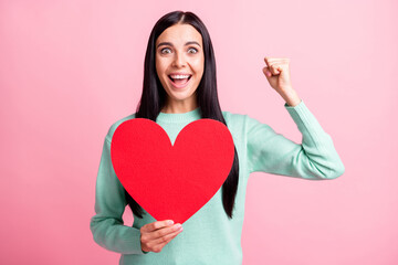 Photo portrait of excited girl celebrating victory with fist up holding big red heart card in one hand isolated on pastel pink colored background