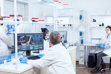 Fototapeta na wymiar Elderly virologist working on computer with virus on display in modern facility. Senior scientist in pharmaceuticals laboratory doing genetic research wearing lab coat with team in the background.