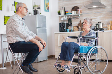 Retired invalid woman in wheelchair having a conversation with old elderly husband in kitchen. Old...