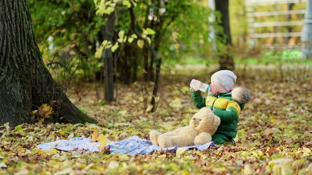 little boy playing with a plush toy in the park. funny baby otty in a warm down jacket and his teddy bear. close up view. Slow motion video. stock footage