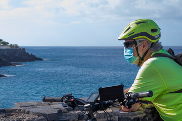 Coronavirus. Portrait of a senior man with bicycle and helmet in sport activity enjoying nature and sea, wearing a protective face mask due to coronavirus contagion