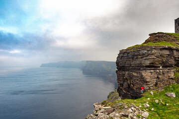 Male tourist in red shirt is climbing stone cliff, Epic view of rock structure of Cliff of Moher in the background, county Clare, Ireland. Cloudy sky.