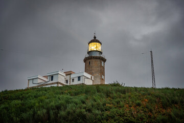 Fototapeta na wymiar old lighthouse on a coastal hill of a small town, the lighthouse has a yellow flashing light, it is night and the sky is dramatic, gray with clouds