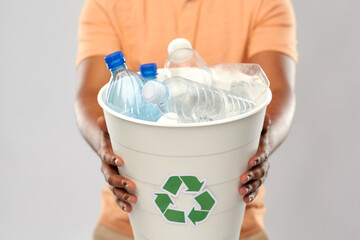 recycling, waste sorting and sustainability concept - close up of african american young man holding bucket with plastic bottles over grey background
