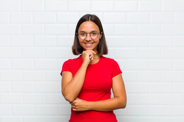latin woman looking happy and smiling with hand on chin, wondering or asking a question, comparing...