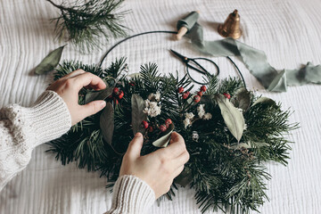 Christmas moody still life. Closeup of women hands crafting advent floral hoop wreath. Garland of fir, eucalyptus. Pine tree branches. Vintage scissors and bell on muslin table cloth. Selective focus.