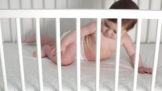 Newborn cheerful caucasian baby is chewing and playing in his crib. Funny smiling looks through the railing of the crib at the camera side view.