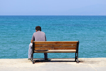 Naklejka na ściany i meble Lonely man sitting on a wooden bench on blue sea background, rear view. Vacation on a beach during coronavirus pandemic