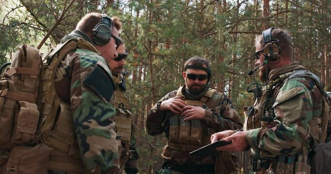 A group of middle-aged bearded soldiers in uniforms and tactical vests discusses the action plan and prepares for action at a temporary forest base. The commander gives instructions to the soldiers