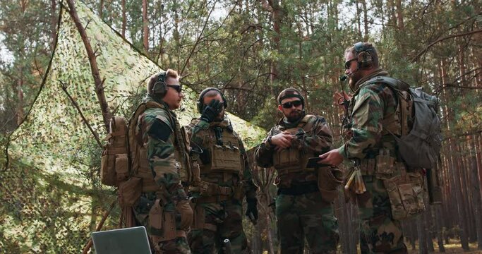 A group of middle-aged bearded soldiers in uniforms and tactical vests discusses the action plan and prepares for action at a temporary forest base. The commander using military tablet.