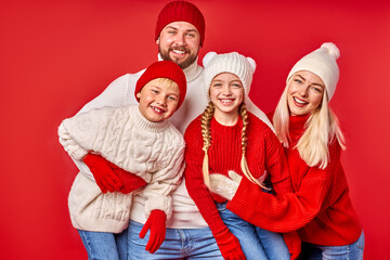 caucasian family in warm winter clothes posing at camera in studio with red background, every member of family in hats, gloves and sweaters, waiting for christmas
