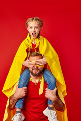 shocked super girl sits on neck of father closing his eyes, superhero man hold child and smiles. kid girl with opened mouth looks at camera isolated over red background