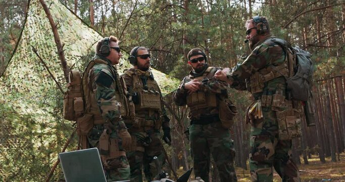 A group of middle-aged bearded soldiers in uniforms and tactical vests discusses the action plan and prepares for action at a temporary forest base. The commander gives instructions to the soldiers.