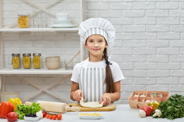 smiling little child girl in cap and an apron knead the dough in the kitchen