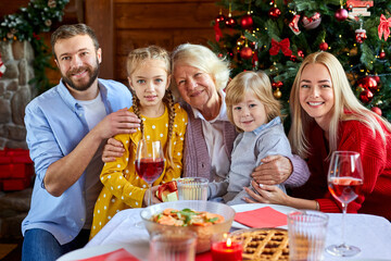 family at home sitting together at table, look and smile at camera, parents with children and grandmother, celebrating christmas
