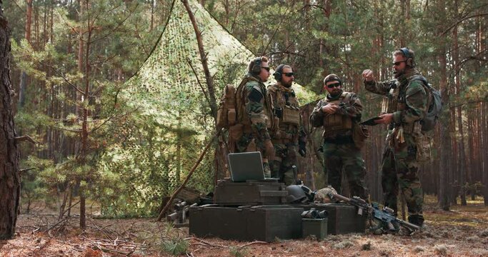 A group of middle-aged bearded soldiers in uniforms and tactical vests discusses the action plan and prepares for action at a temporary forest base. The commander gives instructions to the soldiers