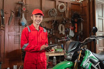 Smiling mechanic wearing wearpack and hat using digital tablet standing near the dirt bike at the garage