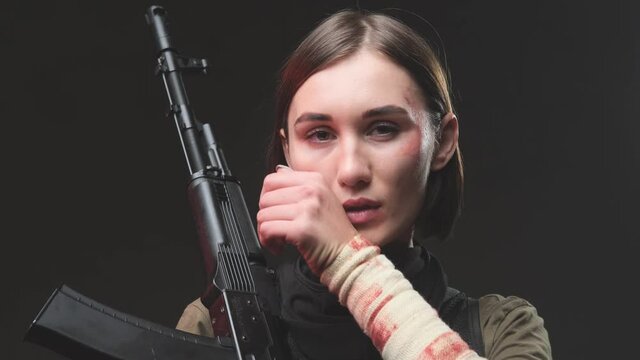 Seductive military woman with short haircut poses in dark background holding a rifle on her shoulder and tucking her hair back behind her ears and then looks at camera.