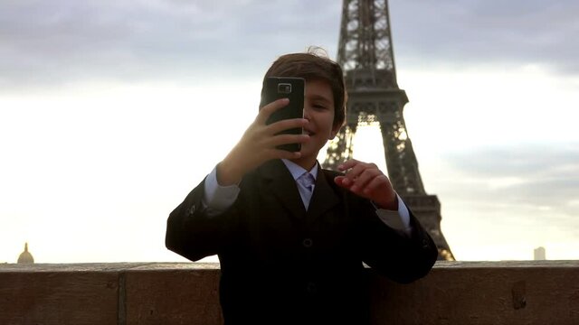 A smiling cute boy in a suit is taking a selfie on the phone on the background of the Eiffel tower at the dawn, Paris, France