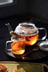 Hot tea with orange and berries in a glass teapot and in a glass mug next to the window in the morning. Sliced orange on a board on a dark background.