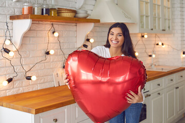 Happy young woman standing in the kitchen, holding huge Valentine balloon and smiling