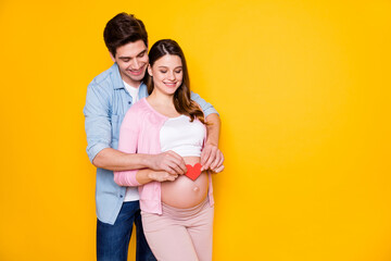 Photo of young couple stand hug pregnant woman show paper heart wear casual outfits isolated on yellow color background