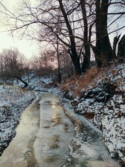 frozen small river with snow capped banks against the backdrop of trees with fallen leaves