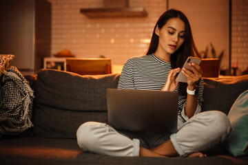 Serious charming woman using cellphone while working with laptop