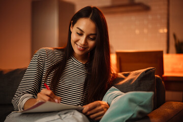 Obraz premium Smiling beautiful woman writing down notes while sitting on sofa