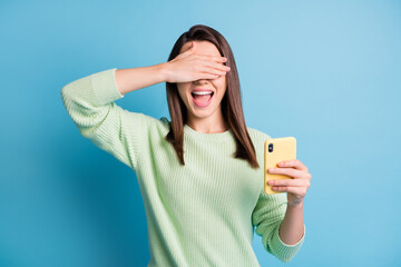 Photo portrait of excited girl with open mouth covering eyes with one hand holding phone isolated on pastel blue colored background