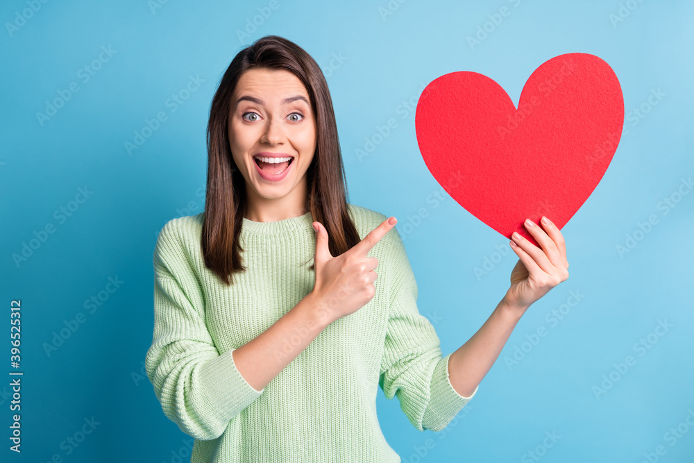 Poster Photo portrait of girl pointing one finger holding big red heart postcard with open mouth isolated on pastel blue colored background