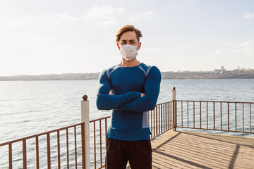 Handsome caucasian male in sportswear poses on the beach wearing a mask. young sportsman male walks outside at sunset by the sea in his protective mask.