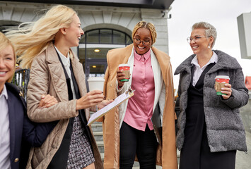 team of diverse business ladies take break outdoors, drink coffee after meeting, leaving building, wearing coats and formal wear. african caucasian cooperation, business concept