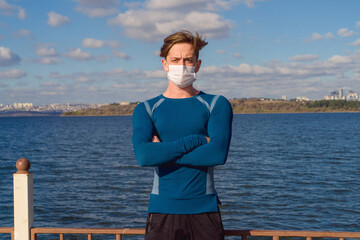 Fototapeta na wymiar Male model posing with mask by the sea on a sunny autumn day. Image of handsome caucasian young man wearing a mask under blue sky with sportswear. Avaible copy space area. 